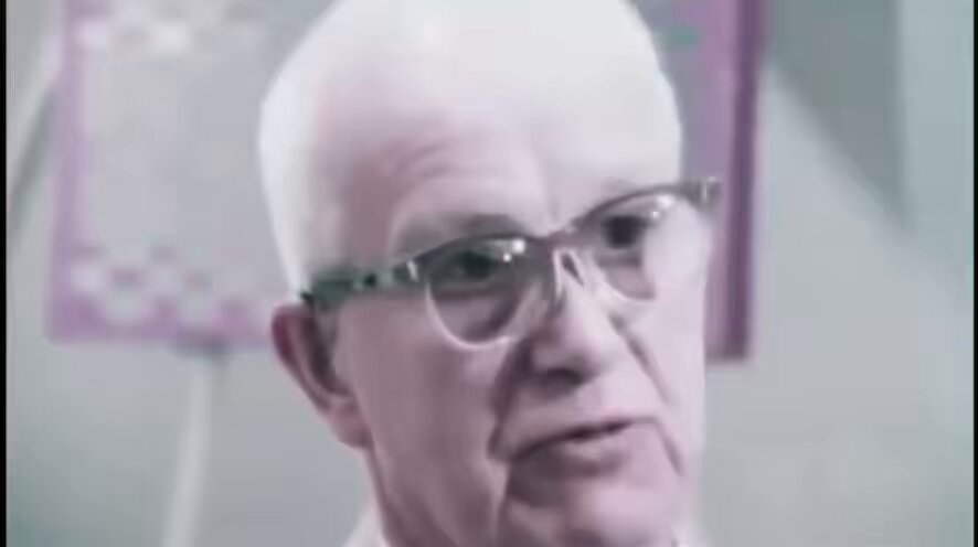 The image features an older man wearing glasses with white hair, who appears to be looking down at something off-camera. The background includes a purple wall and a whiteboard or bulletin board. There are also some words written on the whiteboard that read "Buckminster Fuller predicted Bitcoin in 1967: Energy is wealth". This phrase suggests that the man might be discussing the concept of energy being used to create wealth, which could potentially relate to Bitcoin as an example of digital currency.