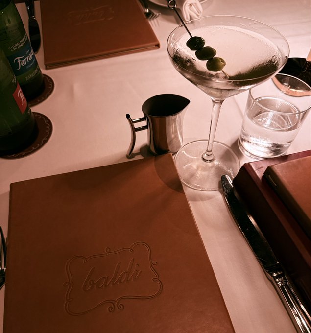 A table setting featuring a martini glass with olives, a water glass, a small pitcher, a green Ferrarelle water bottle, and menus with "baldi" embossed on them.