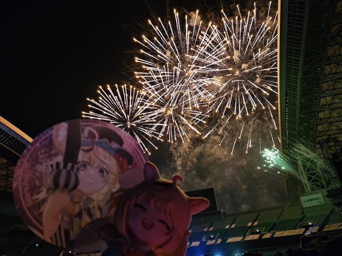 Bright fireworks burst in the night sky above a building, with an anime-themed inflatable balloon featuring two characters in the foreground.