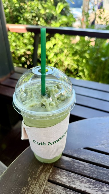 A green smoothie or frozen drink from Café Amazon in a clear plastic cup with green straw, sitting on a wooden table outdoors.