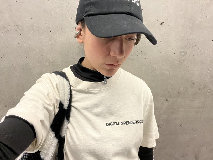 Selfie of a young woman wearing a black cap and a white sweatshirt with 'DIGITAL SPENDERS GU...' text printed on it, standing against a concrete wall.
