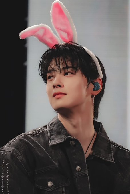 A young man with dark hair wearing pink and white bunny ears and a denim jacket looks upward, with a blue in-ear monitor visible.