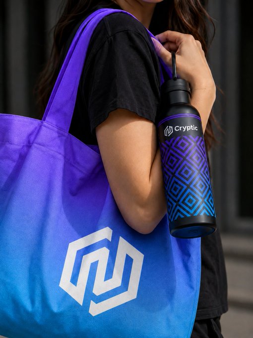 A person carries a purple-to-blue gradient tote bag featuring a white Cryptic logo alongside a matching black water bottle labeled "Cryptic" with a geometric blue design, showcasing the brand's coordinated lifestyle accessories.
