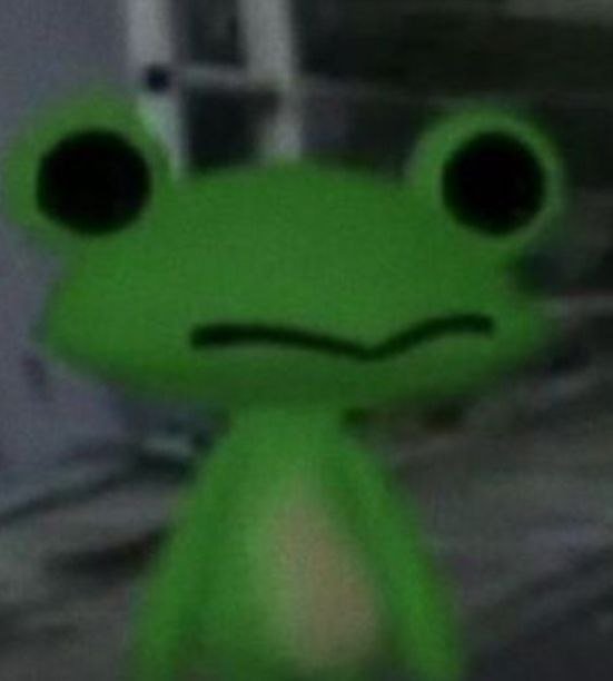 A low-resolution, blurry close-up image of a cartoon-like green frog character with large black eyes and a subtle smile, set against a dark, indistinct background.
