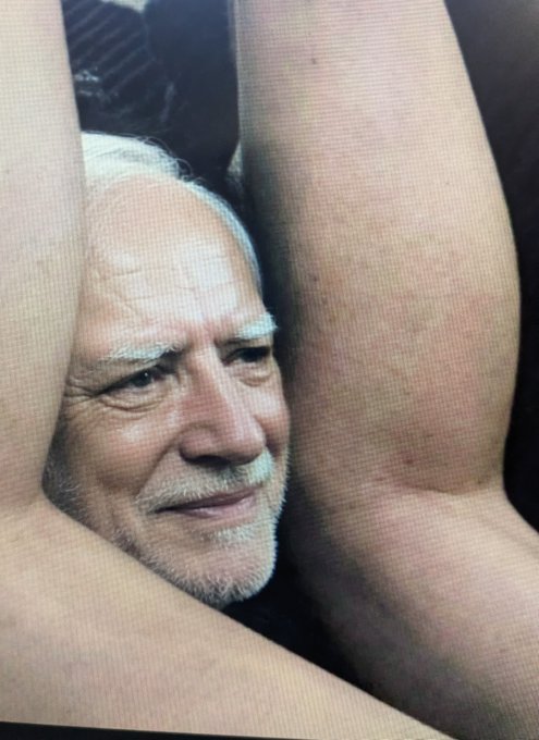 A photograph of a TV screen displaying a close-up of an older man's face squeezed between a person's legs in a sexually suggestive pose.