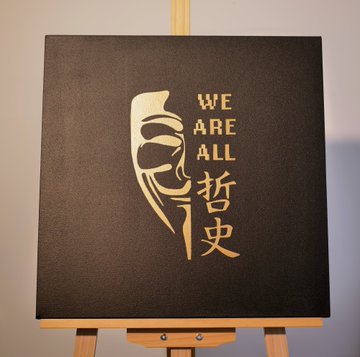 A black canvas on an easel featuring a gold silhouette of a masked figure alongside the text 'WE ARE ALL 哲史'.