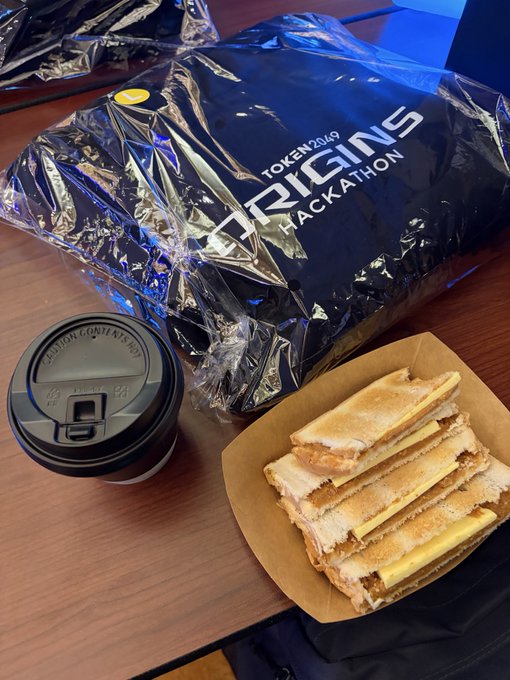 A black 'ORIGINS HACKATHON TOKEN2049' bag sits next to a coffee cup and a plate with a grilled cheese sandwich on a wooden table.