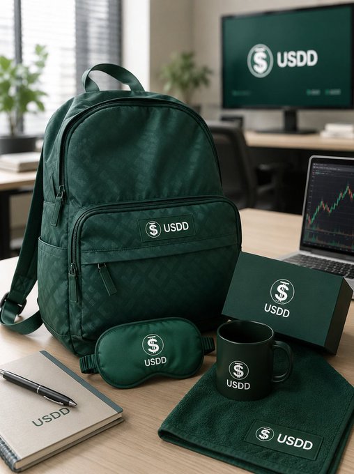 A collection of dark green branded merchandise including a backpack, notebook, mug, eye mask, and towels, all featuring the "USDD" logo and emblem, arranged on a desk with a computer monitor.