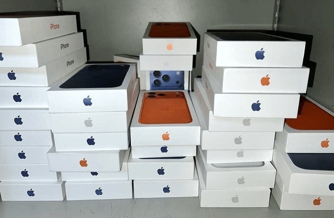 A stack of sealed iPhone boxes featuring the Apple logo, with one box open revealing a blue iPhone camera module, tied to a giveaway contest where 20 random winners will be selected from numbers 20–72 to win a $5,000 surprise.