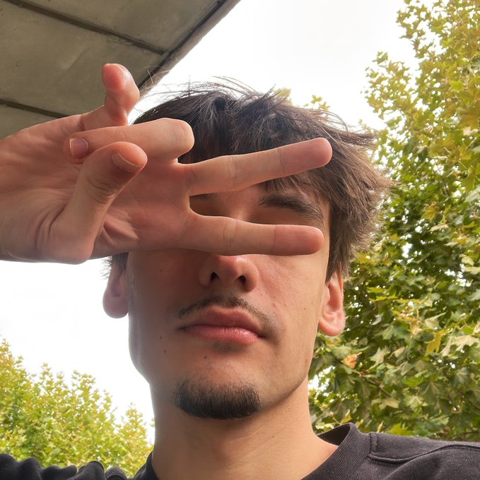 A young man with tousled dark hair and a goatee holds up three fingers in a casual, low-angle selfie, partially shielding his face from the bright sky as he poses outdoors near some trees.
