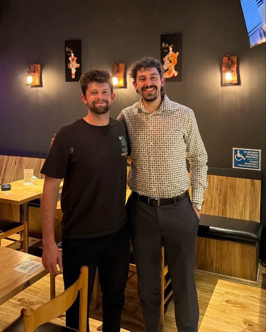 Photo of two men smiling and posing together in what appears to be a restaurant with wood paneling and Asian-style wall decorations.