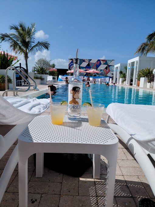 Two margaritas with lime garnishes sit on a white mesh table beside a bottle of "BORED" vodka featuring a cartoon ape character, with a sunny pool scene and palm trees in the background. The ape-themed bottle directly ties to the "Bored Ape" reference in the post, capturing the Miami poolside vibes being celebrated.