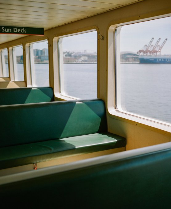 Empty green benches line the "Sun Deck" of a ferry, with large windows revealing a harbor view of industrial cranes and a docked ship. The image captures the quiet, sunlit interior that inspired the sold first edition print, which was just collected by @osseneth as part of the limited 19/20 remaining.