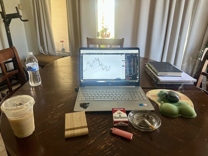 A laptop displaying a financial chart sits on a wooden table alongside a pack of Marlboro cigarettes, an ashtray with a cigarette, a cold drink, and a water bottle.