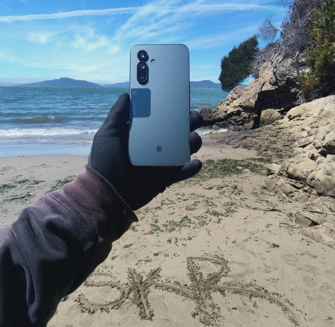 A first-person view of a gloved hand holding a light blue smartphone on a beach, with the ocean and mountains in the background, and Japanese characters written in the sand.