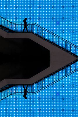 A silhouetted figure walks along a zigzag staircase against a vibrant blue grid background, with their reflection visible on the lower portion of the stairs. The pixelated blue backdrop and minimalist composition reflect the digital NFT artwork "Endless Blue" by gulyildizart, recently acquired on objktcom.