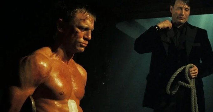 Two men stand in a dimly lit room; a shirtless, sweating man with a bandage on his side appears battered on the left, while a man in a black suit holding a coiled rope stands on the right.