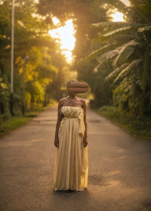 A woman in a flowing yellow gown stands on a tree-lined road bathed in golden sunset light, her face obscured by a surreal wooden, hollowed form—evoking the artwork's theme of unseen inner selves, as referenced in the accompanying caption about hidden parts of identity.