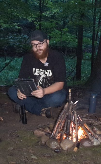 A bearded man in glasses and a 'LEGEND' t-shirt sits by a campfire in the woods, looking down at a tablet in his hands.