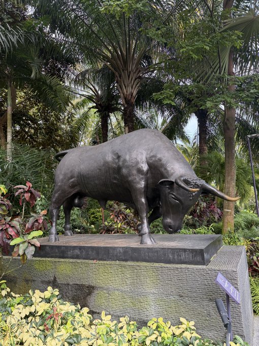 A dark metal statue of a bull with long horns standing on a stone pedestal, surrounded by tropical palm trees and foliage.