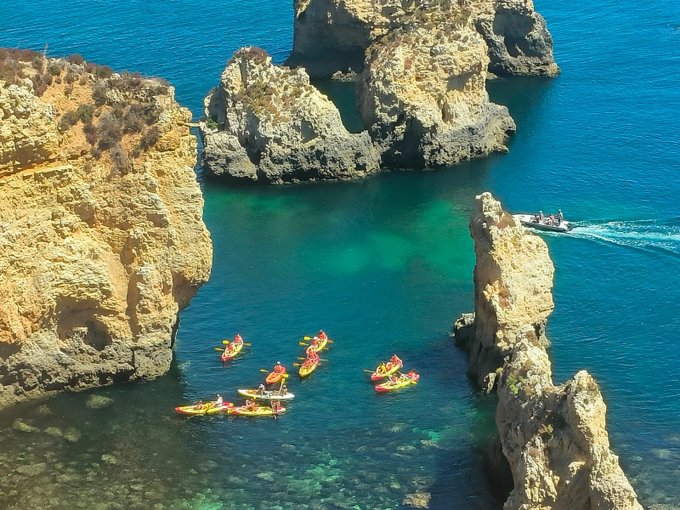 An aerial view shows a group of kayakers paddling through crystal-clear turquoise waters between dramatic golden rock formations at the coast.