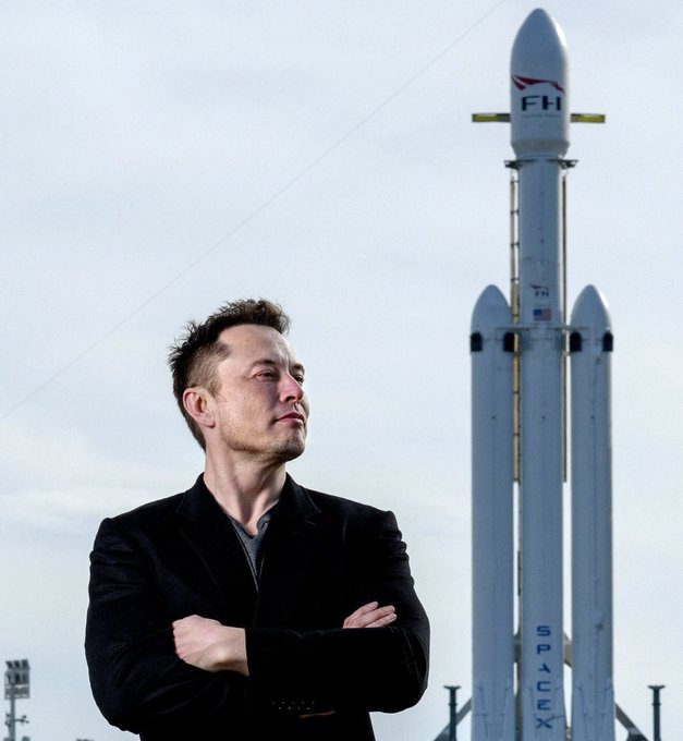 Elon Musk stands with arms crossed in a black jacket in front of a SpaceX Falcon Heavy rocket on the launch pad under an overcast sky.