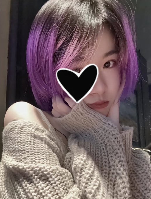 A selfie of a young woman with purple hair wearing a cream-colored knit sweater, with a black heart obscuring one of her eyes.
