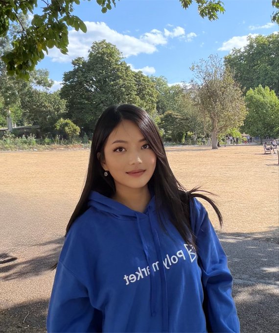 A young woman with long dark hair wearing a blue 'Body of the Artist' hoodie, posing in a park with trees and blue sky in the background.
