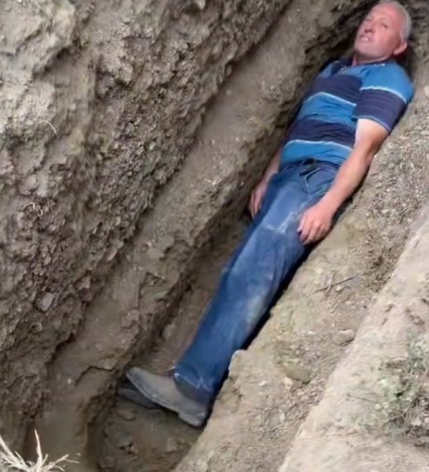 The image shows a man lying down in a hole or trench filled with dirt. He appears to be resting or sleeping, as he has his legs crossed over each other. The surrounding area seems to be a construction site or an excavation pit. There is no visible text related to the price of ETH on this image.