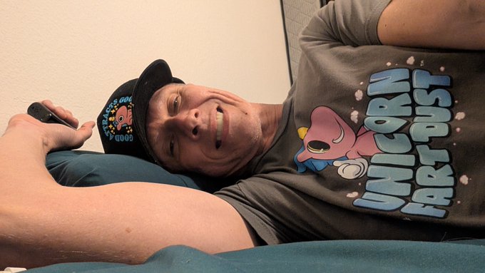 A man wearing a black "Unicorn Fart Dust" cap and gray t-shirt lies back on a couch, smiling while holding a remote, as he continues his day 558 streak of wearing the shirt to promote UFD.
