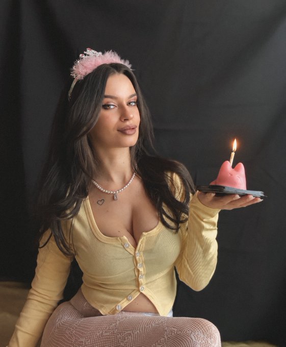 A woman wearing a pink fluffy headband and a yellow cardigan holds a small plate with a lit pink candle, posed against a black backdrop.