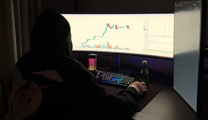 A hooded figure sits at a desk with a colorful RGB keyboard, analyzing a stock trading chart on a large monitor in a dark room, with a green bottle nearby.