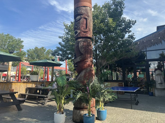 A large carved wooden tiki-style totem pole stands at the center of an outdoor patio area, surrounded by potted palm plants, picnic tables under umbrellas, and what appears to be a ping pong table, suggesting a tropical-themed bar or restaurant. The casual seating and recreational setup fit the relaxed social vibe of the post, which hints at relocating for activities like Drink and Draw or Karaoke.