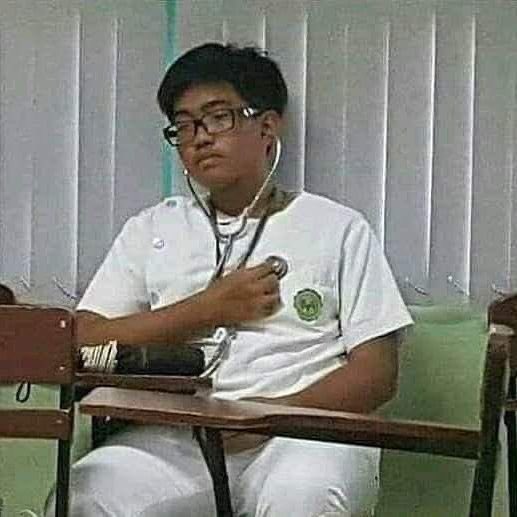 A young man in a white school uniform with glasses and a stethoscope around his neck, appearing to listen to his own chest in a classroom.