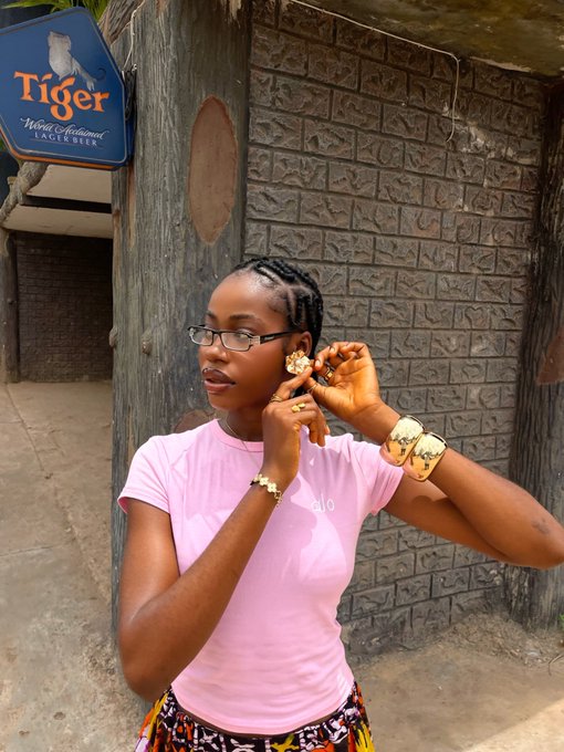A young woman wearing glasses and a pink t-shirt with gold jewelry, posing in front of a 'Tiger' Lager Beer sign and a brick wall.