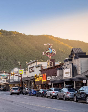 A scenic photograph of a small mountain town street with parked cars, various storefronts, and forested hills in the background during golden hour.