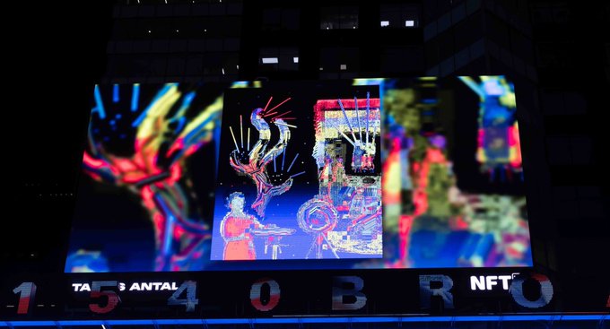 Nighttime photo of a building facade displaying 'TARSANTAI 4 0 BRO NFT' below a vibrant, colorful digital art projection featuring abstract figures.