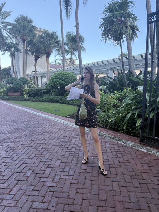 A woman in a black floral dress and sandals stands on a brick pathway holding a piece of paper and a small gold bag, surrounded by palm trees and tropical landscaping.