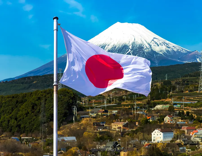 A photograph of the Japanese flag (white with a red circle) waving in the foreground, with snow-capped Mount Fuji and a small town visible in the background under a clear blue sky.