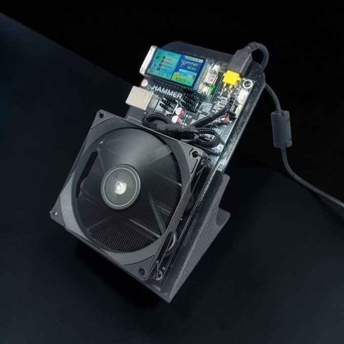 The image shows a Hammer Miner BC04, a compact desktop Bitcoin mining device featuring a cooling fan, a circuit board, and a small display screen, mounted on a dark base with a power cable. The branding "HAMMER MINER" is visible on the device, which corresponds to the 6 TH/s home Bitcoin miner mentioned in the post, designed for casual home cryptocurrency mining.