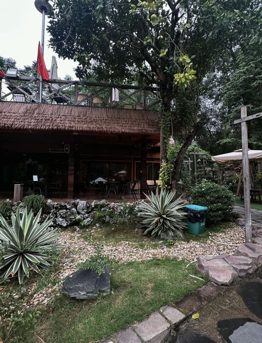 An outdoor garden scene featuring a thatched-roof building, tropical plants, a large tree, and a stone-lined pond with stepping stones.