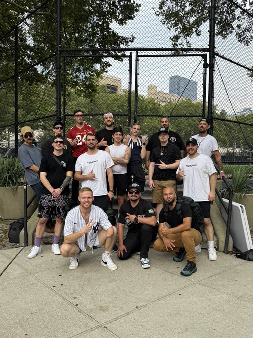A large group of around 14 men pose for a group photo in front of a chain-link fence, with city buildings and trees visible in the background.