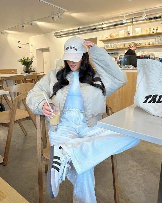 The image features a woman sitting at a table, wearing a beige jacket and white pants while holding a drink. She has her hair styled in pigtails and appears to be enjoying her time. The background of the photo shows several chairs placed around tables, suggesting that she is in a cafe or similar establishment. The caption for this image reads: "Why am I bullish on @TheARCTERMINAL Because crypto is becoming too complex for the average user. More chains. More protocols. More data. More decisions.ARC Terminal's vision is to simplify all of that with an AI-powered command center where users can understand what's".