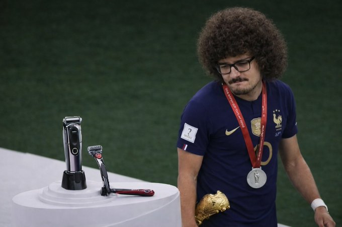 A dejected soccer player with curly hair and glasses, wearing a France national team jersey with a silver medal, walks past a pedestal holding the World Cup trophy and a razor.