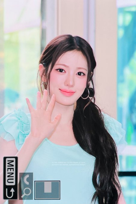 A photo of a young woman with long dark hair wearing a light blue ruffled top, waving at the camera with a gentle smile.