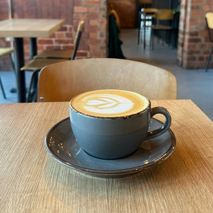 A grey ceramic cup filled with a latte featuring leaf latte art sits on a matching saucer on a light wooden table, with a brick-walled cafe interior and other tables visible in the background.