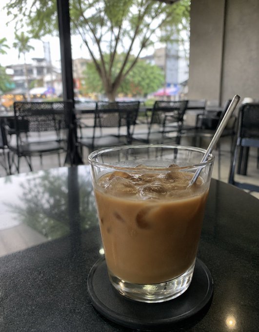 A glass of iced coffee with milk sits on a dark coaster on a table, with a metal straw, looking out toward an outdoor patio with chairs and a tree.
