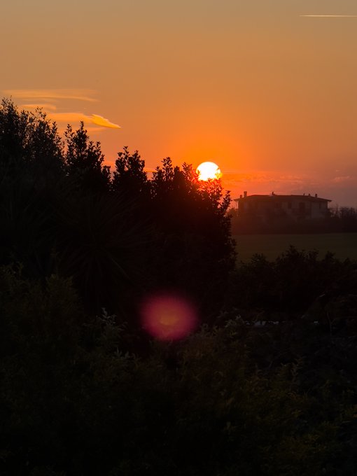 The image features a beautiful sunset over trees, with the sun setting behind the trees casting a warm glow across the sky. The scene includes a house visible in the background, adding depth and context to the photograph. The photo has been shared on social media platforms, where it is being appreciated by many users for its artistic composition and stunning visual appeal.