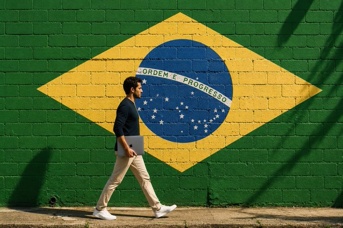 A man with a laptop walks past a brick wall painted with the Brazilian flag, featuring the iconic green, yellow, and blue colors and the motto 'ORDEM E PROGRESSO'.