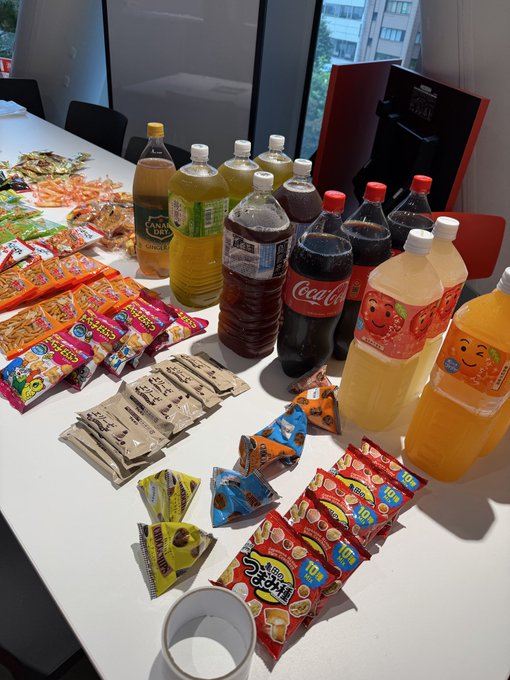 A table is filled with an assortment of snacks and beverages, including Coca-Cola bottles, orange juice, tea, seaweed snacks, and various colorful snack packages, with an empty white cup in the foreground.